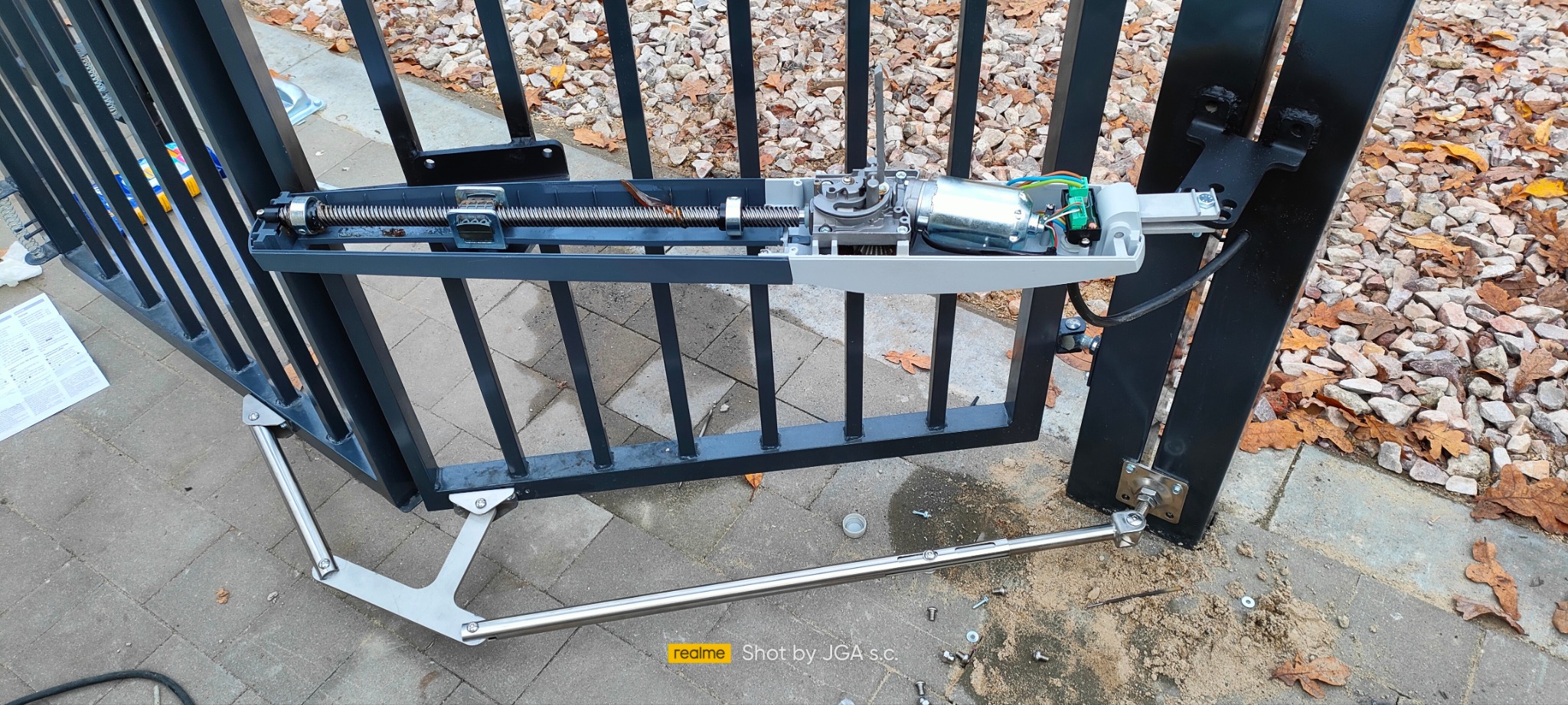 Widok z bliska na zamontowany mechanizm automatyki bramy skrzydłowej, z widocznymi elementami silnika, przekładni ślimakowej i ramienia teleskopowego, na tle kostki brukowej i opadłych liści.