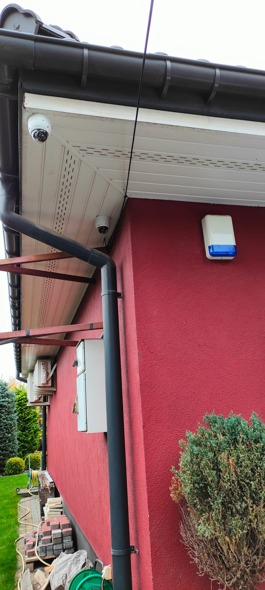 Dwie kamery kopułkowe zamontowane pod okapem czerwonego domu, obok rynna spustowa i skrzynka elektryczna, widoczny również sygnalizator alarmowy z niebieskim paskiem.