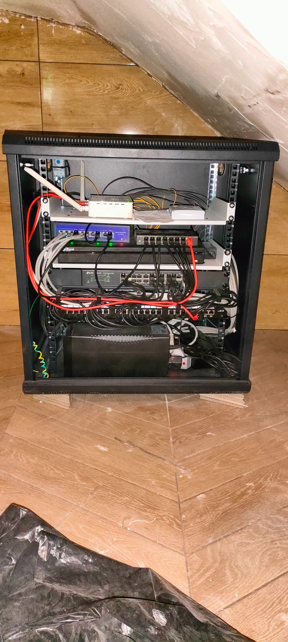 Czarna szafa rack z zainstalowanymi urządzeniami sieciowymi, w tym routerem, switchem i UPS, umieszczona na drewnianej podłodze w pomieszczeniu ze skośnym sufitem.