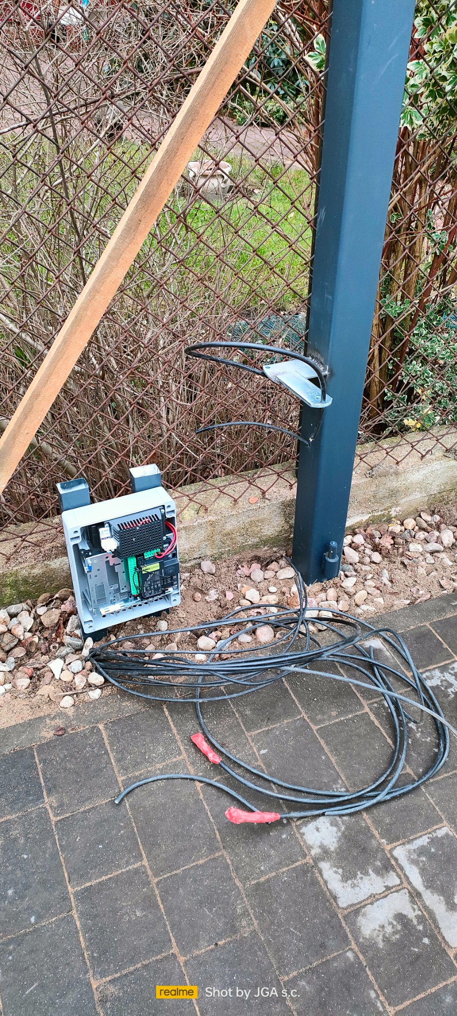 Otwarta skrzynka sterownicza z widocznymi elementami elektroniki, kable wychodzące na zewnątrz, zamontowana na zewnątrz, obok słupka ogrodzeniowego z uchwytem na antenę.