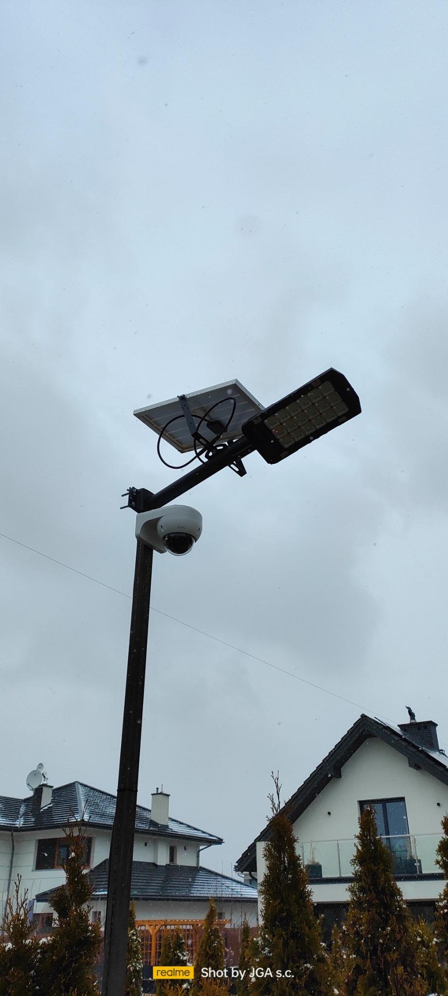 Słup oświetleniowy z panelem solarnym i kamerą monitoringu na tle domów w zimowej scenerii.
