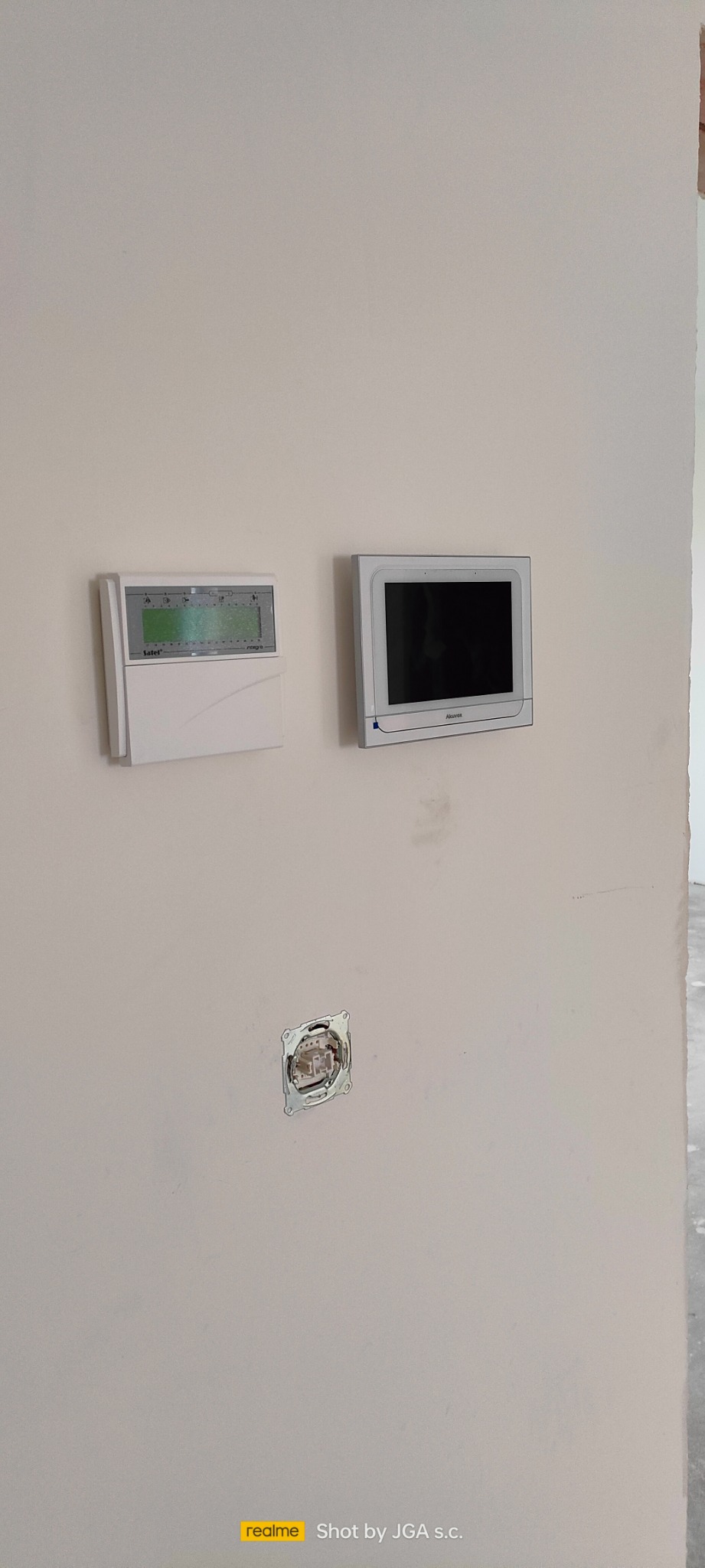Panel alarmowy Satel Integra i wideodomofon Akuvox zamontowane na ścianie, widoczne gniazdko elektryczne bez osprzętu.