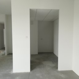 Osoba prywatna - Wnętrze w stanie deweloperskim z surową podłogą i świeżo pomalowanymi na biało ścianami, widoczny fragment wiadra z farbą w tle.