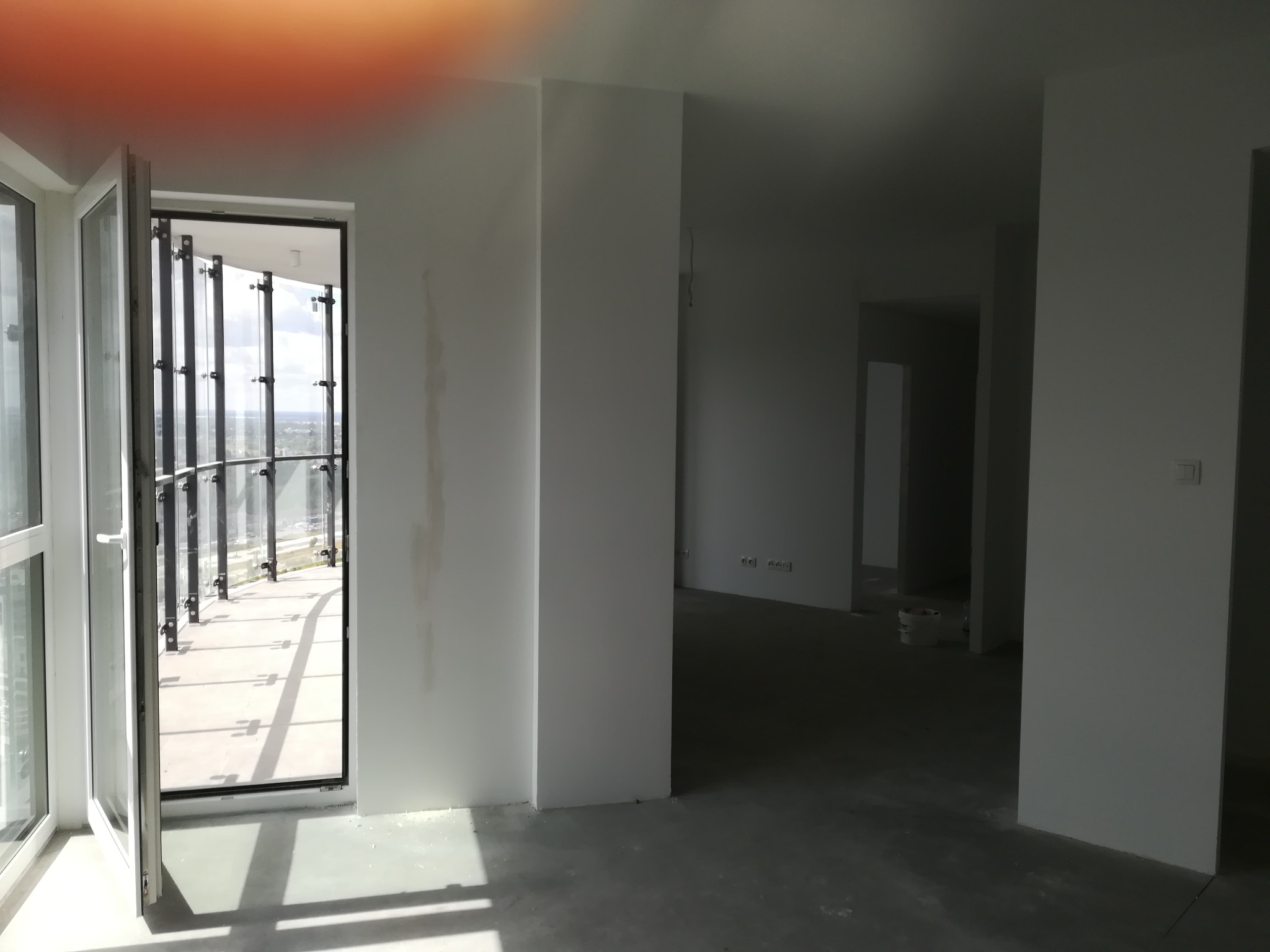 Surowe wnętrze apartamentu w trakcie wykańczania, widoczne świeżo pomalowane ściany na biało, otwarte drzwi balkonowe z widokiem na miasto i szklaną balustradę balkonu, surowa betonowa podłoga...