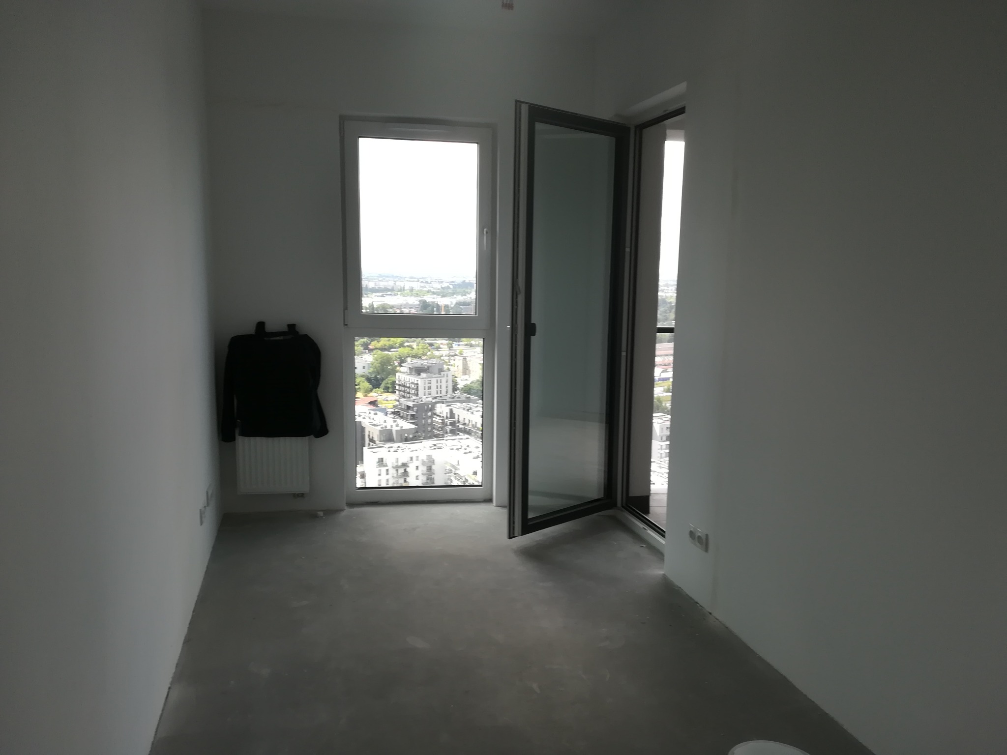 Puste, jasne pomieszczenie w stanie deweloperskim z widokiem na miasto przez okno i otwarte drzwi balkonowe; czarna kurtka wisząca na grzejniku.