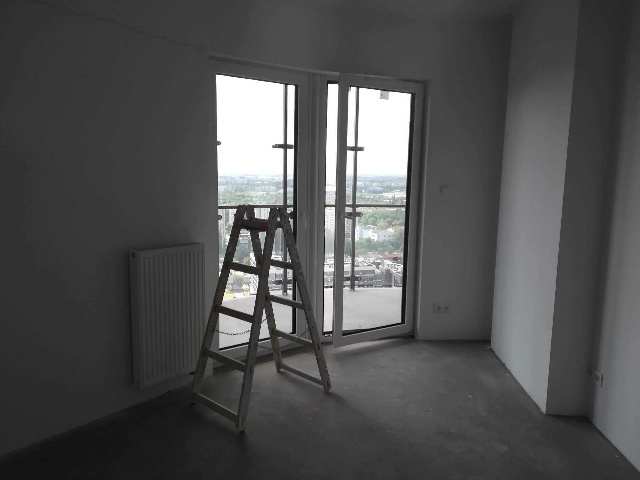 Pusty pokój z grzejnikiem i drabiną przed oknem balkonowym z widokiem na miasto, przygotowany do prac wykończeniowych.