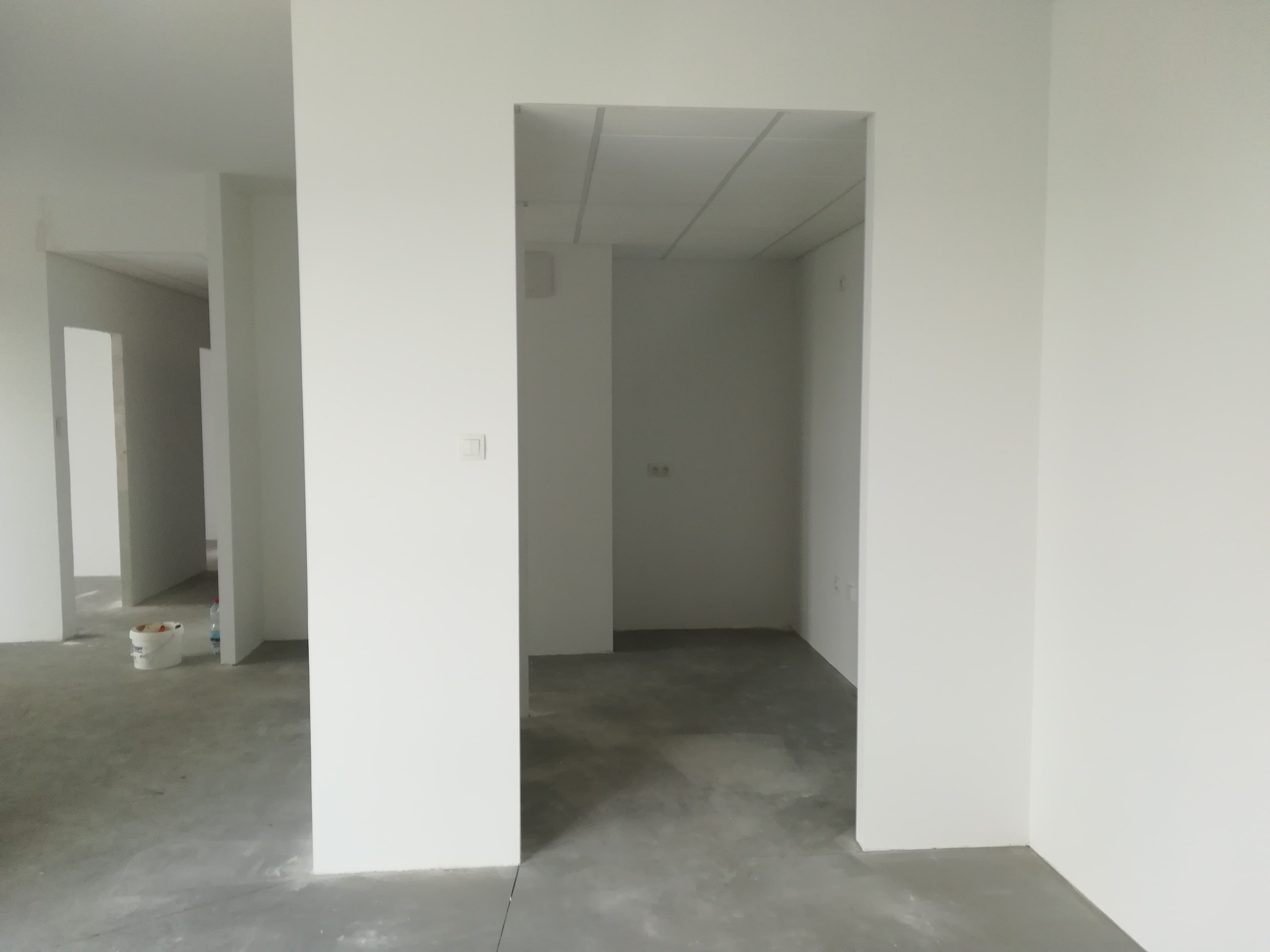 Wnętrze w stanie deweloperskim z surową podłogą i świeżo pomalowanymi na biało ścianami, widoczny fragment wiadra z farbą w tle.