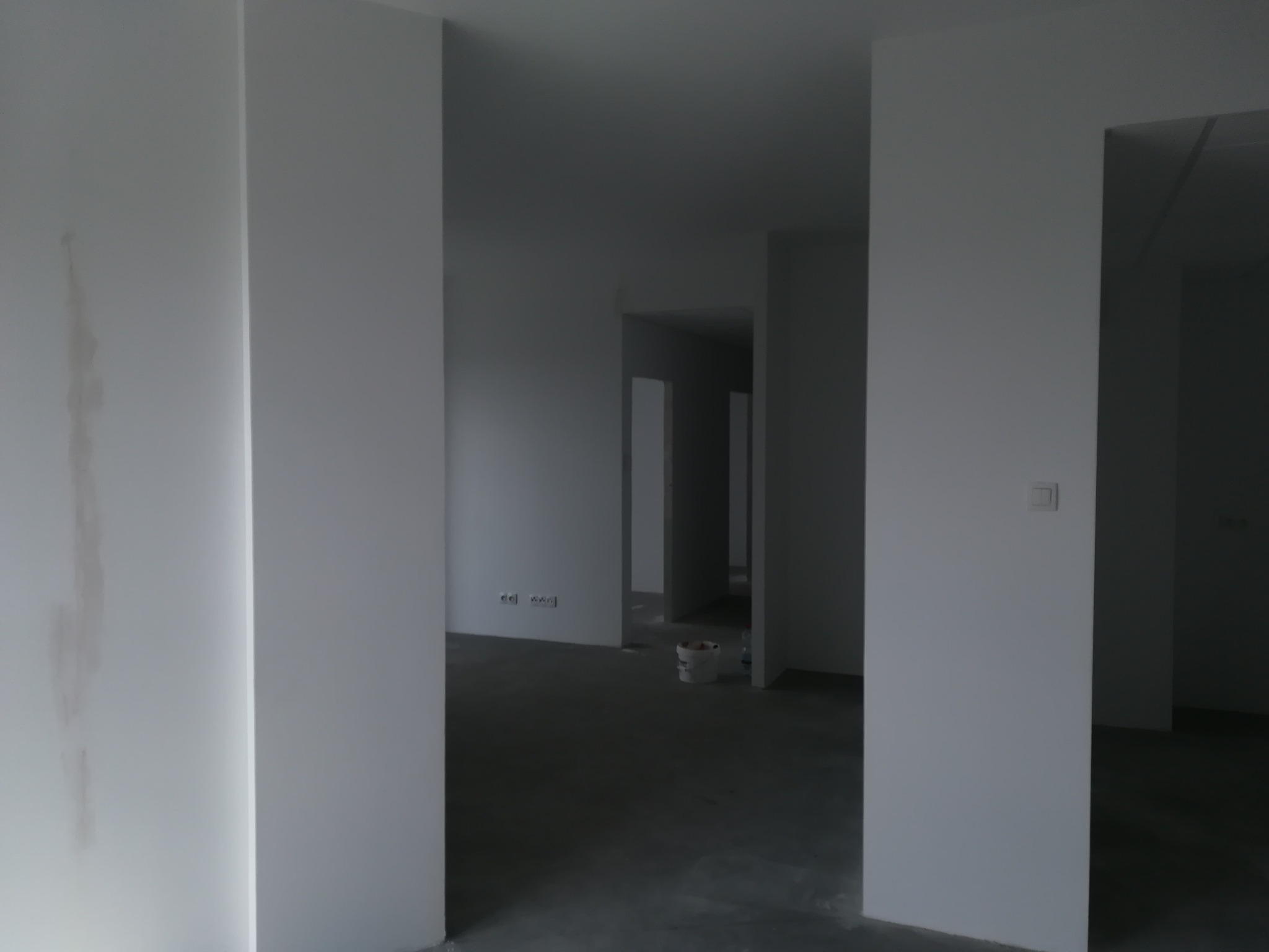 Wnętrze w trakcie przygotowań do prac wykończeniowych, widoczne ślady po zalaniu na ścianie, surowa podłoga, puszka z farbą w tle, widoczne gniazdka elektryczne.