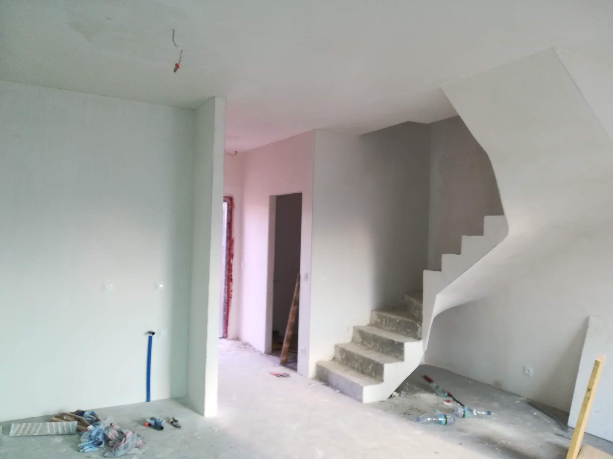 Surowe wnętrze w trakcie prac: świeżo otynkowane ściany, betonowe schody bez wykończenia, wystające przewody i nieporządek na podłodze.