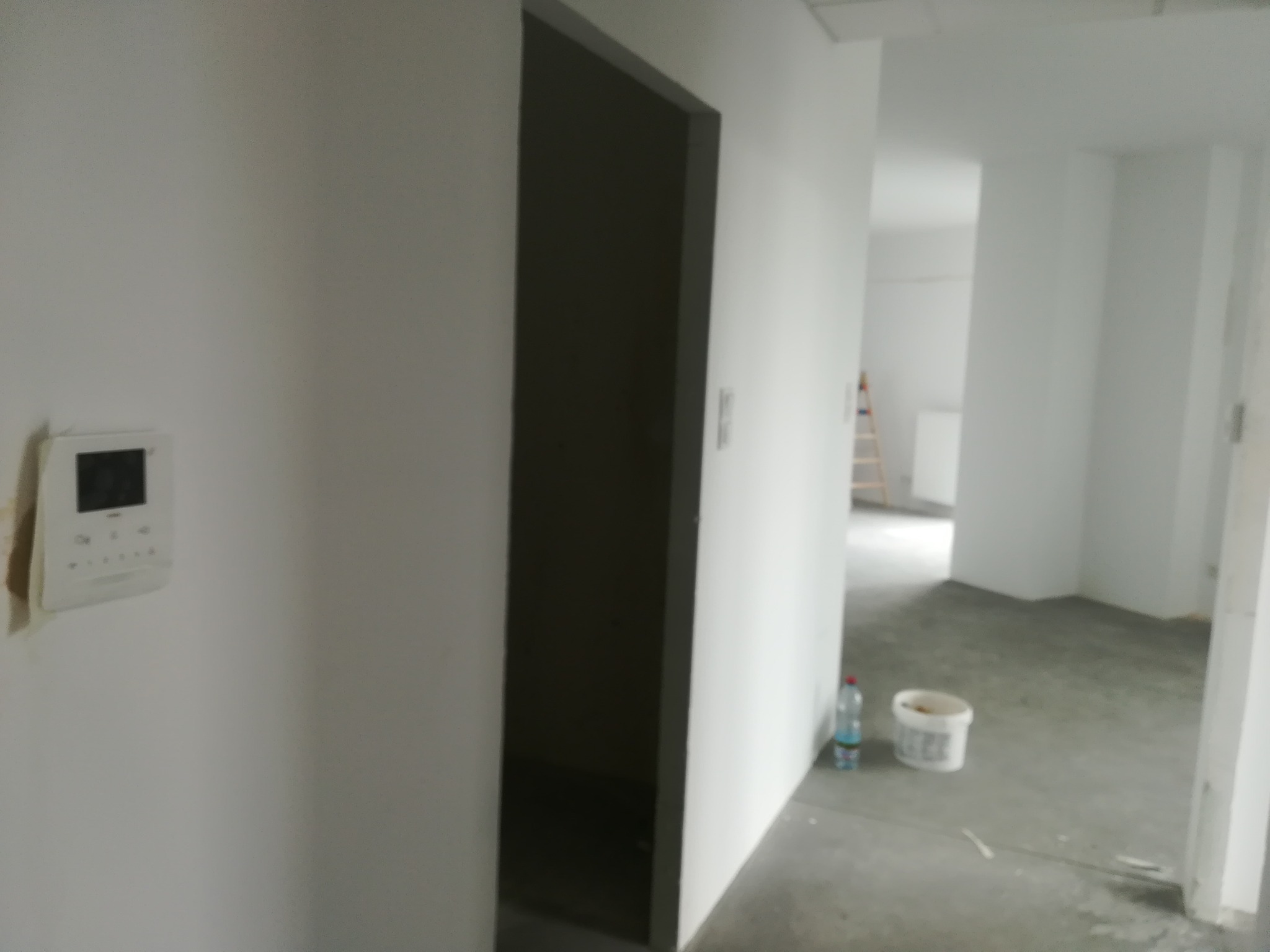Wnętrze w trakcie prac przygotowawczych, widoczne surowe, białe ściany, ciemna ościeżnica drzwi, regulator temperatury na ścianie, wiadro z farbą i butelka wody na podłodze, w tle drabina malarska.