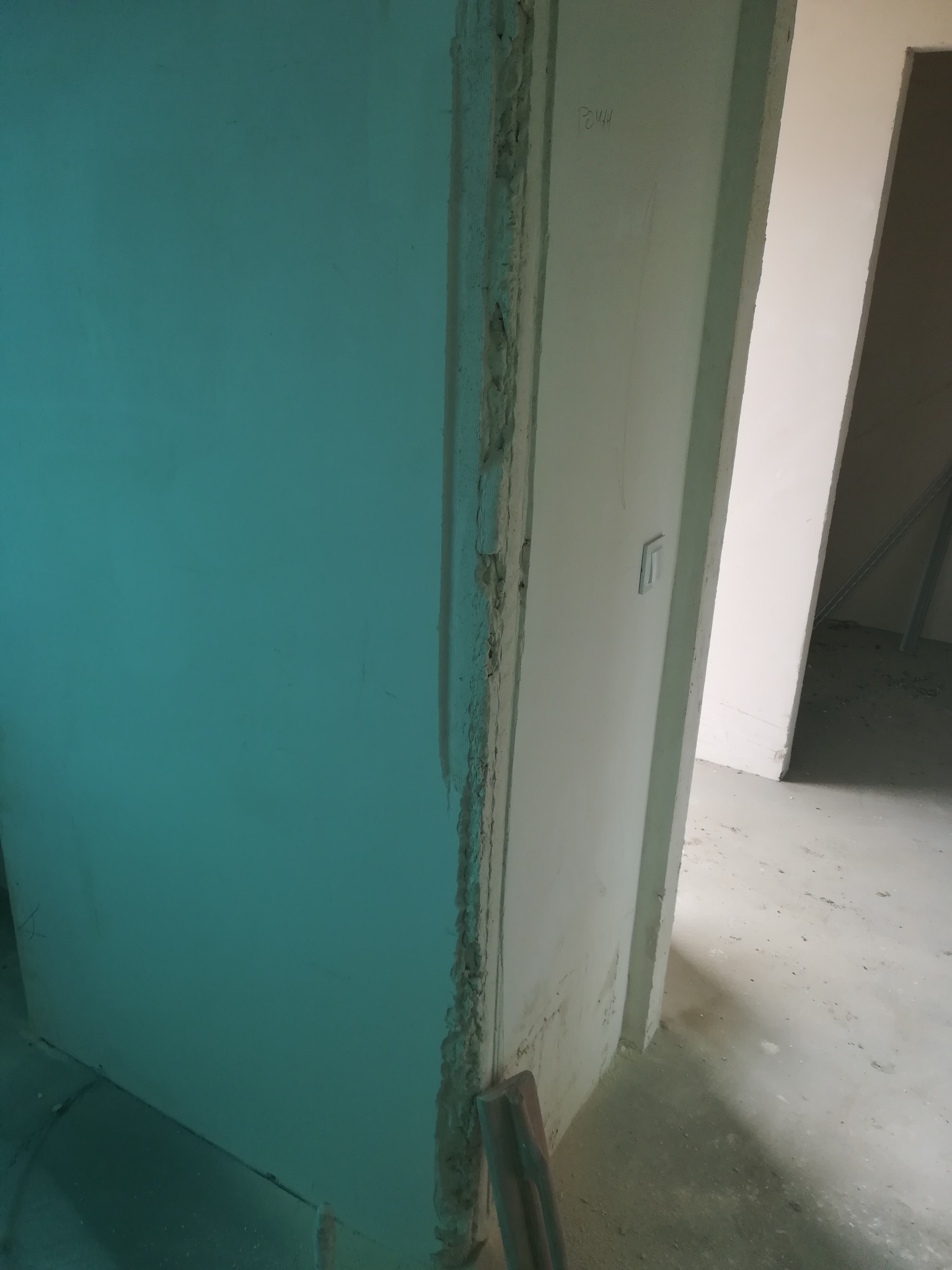 Niedokończona ściana w kolorze turkusowym z widocznymi śladami po szpachlowaniu przy ościeżnicy, widok na sąsiednie pomieszczenie z surową podłogą.