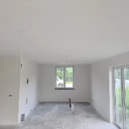 Wnętrze nowo wykończonego pomieszczenia z białymi ścianami, widoczne instalacje elektryczne wystające z sufitu i ścian, surowy betonowy podkład, okno z widokiem na zieleń i częściowo widoczny...