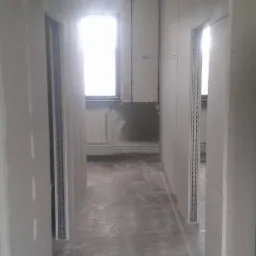 Korytarz w trakcie montażu płyt kartonowo-gipsowych, widoczne profile i szpachlowane łączenia, surowy stan wykończenia z widocznym grzejnikiem i instalacją elektryczną.