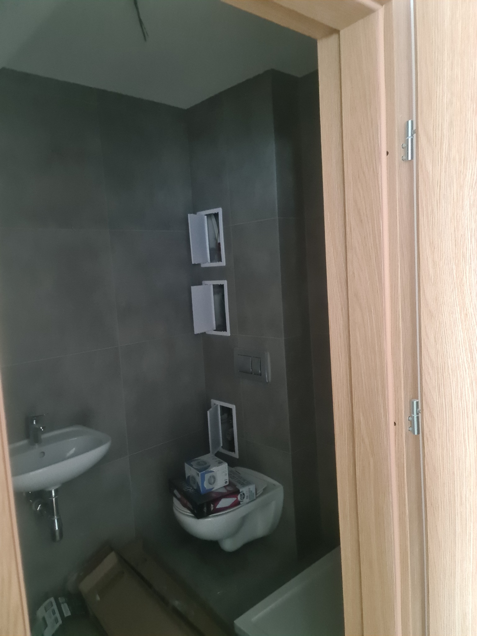 Łazienka w trakcie remontu: szare płytki ścienne, biała umywalka i toaleta z pudełkami na desce, widoczne otwarte drzwiczki rewizyjne i fragment ościeżnicy.