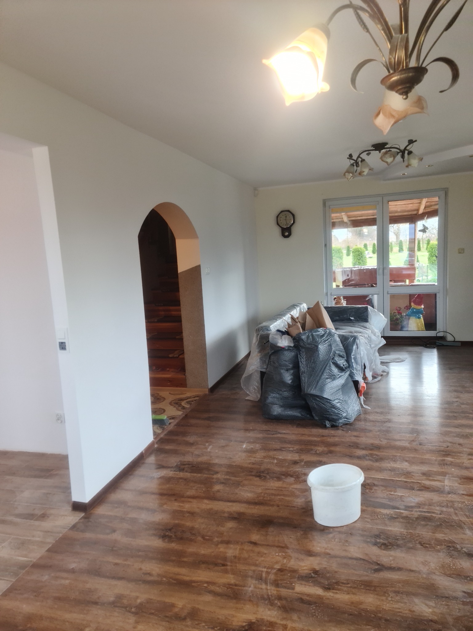 Pokój w trakcie prac wykończeniowych: ściany po malowaniu, zabezpieczone meble folią, widoczne worki z odpadami i wiadro na podłodze z panelami imitującymi drewno.