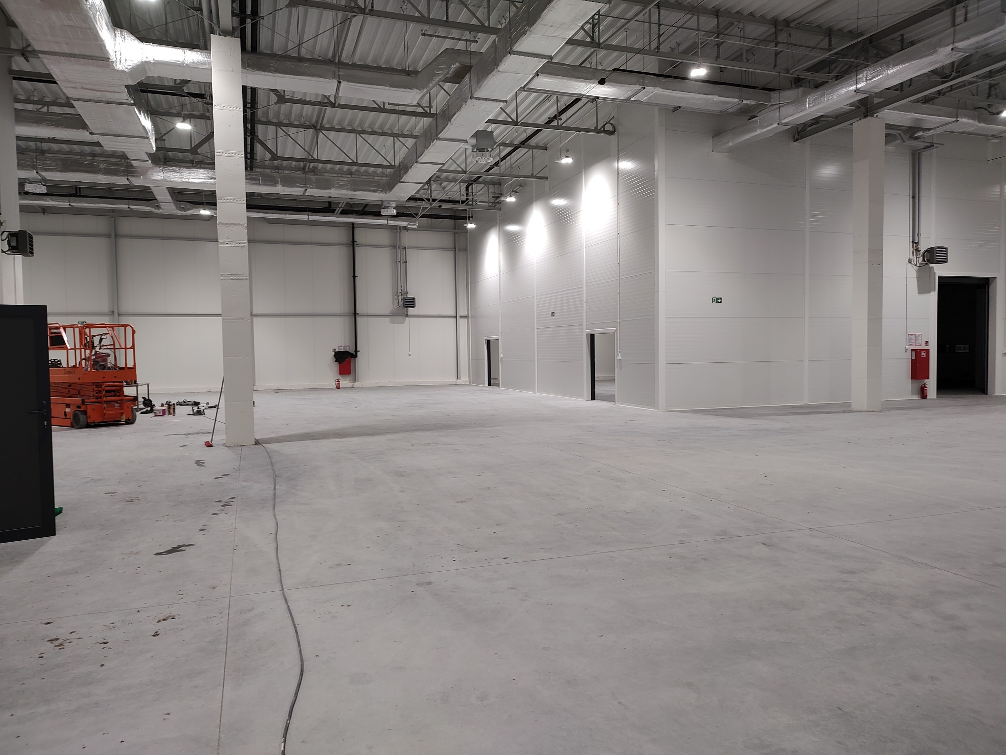 Wnętrze hali przemysłowej, widoczna podłoga z betonu, białe ściany z panelami, instalacje pod sufitem, pomarańczowy podnośnik nożycowy w lewym rogu, ślady na podłodze.