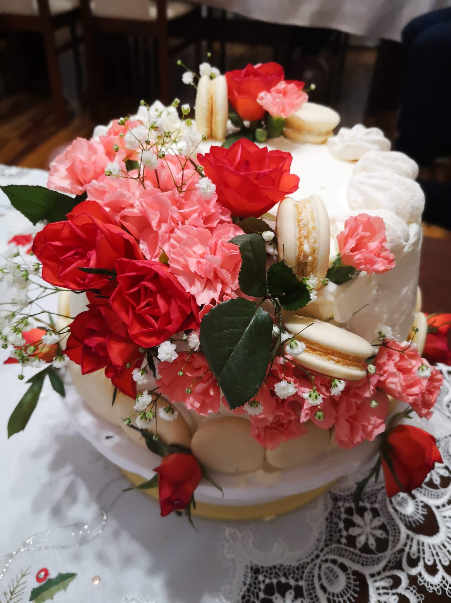 Dwupoziomowy tort śmietankowy udekorowany czerwonymi różami, różowymi goździkami, gipsówką i makaronikami w kolorze ecru, na białym obrusie z koronką.