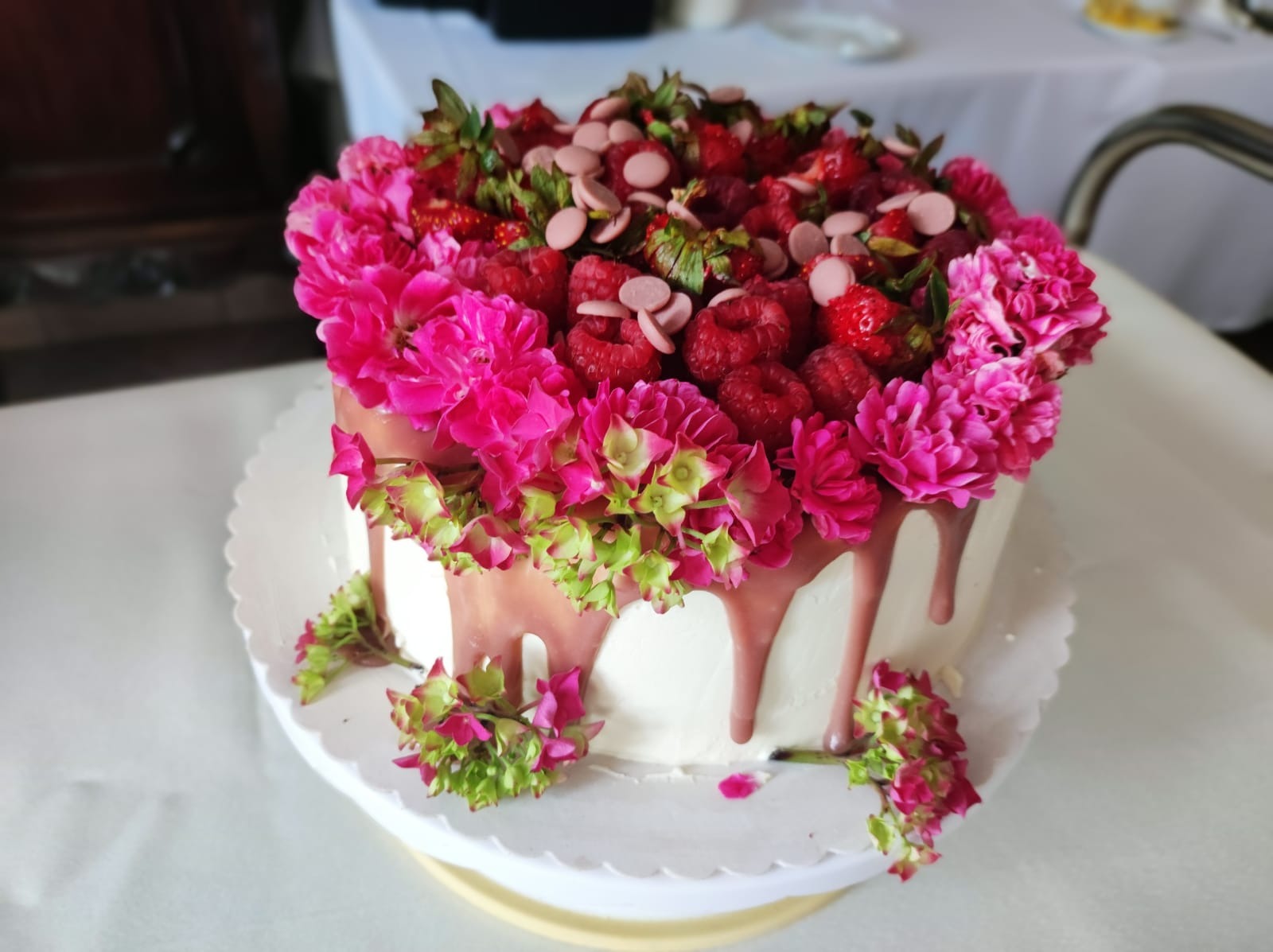 Tort z białym kremem, dekorowany świeżymi malinami, truskawkami, różowymi kwiatami i polewą w kolorze różowym.