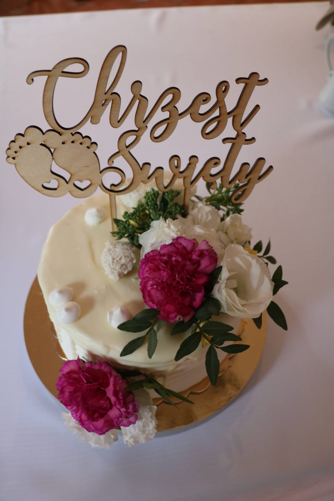 Tort z kremem, dekoracją z żywych kwiatów i drewnianym napisem 'Chrzest Święty' z motywem stópek dziecka na złotej podkładce.