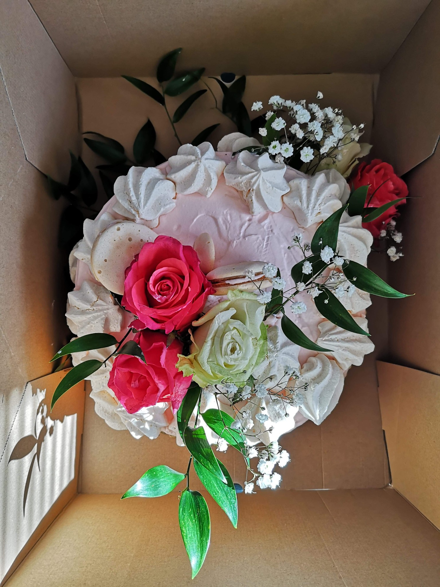 Tort w kartonowym pudełku, udekorowany różowymi różami, białymi bezikami i zielonymi liśćmi, widziany z góry.
