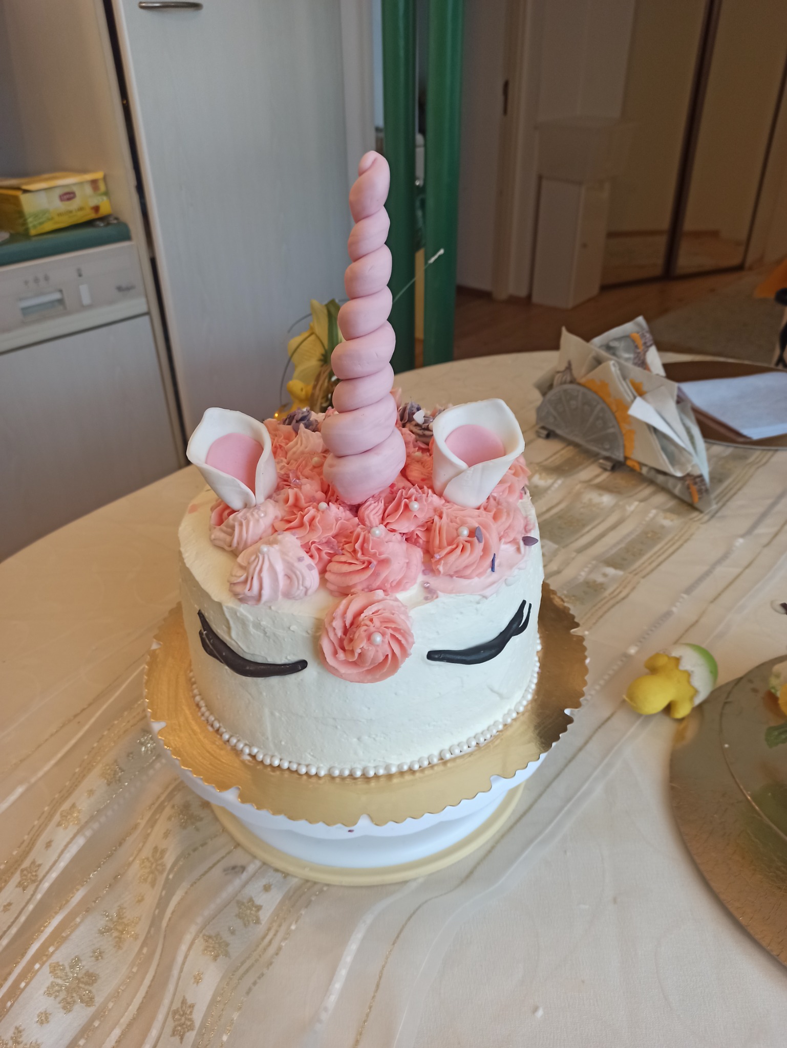 Tort jednorożec z różowymi różami z kremu, białym lukrem, czarnymi rzęsami i rogiem z różowej masy cukrowej, umieszczony na złotej paterze.