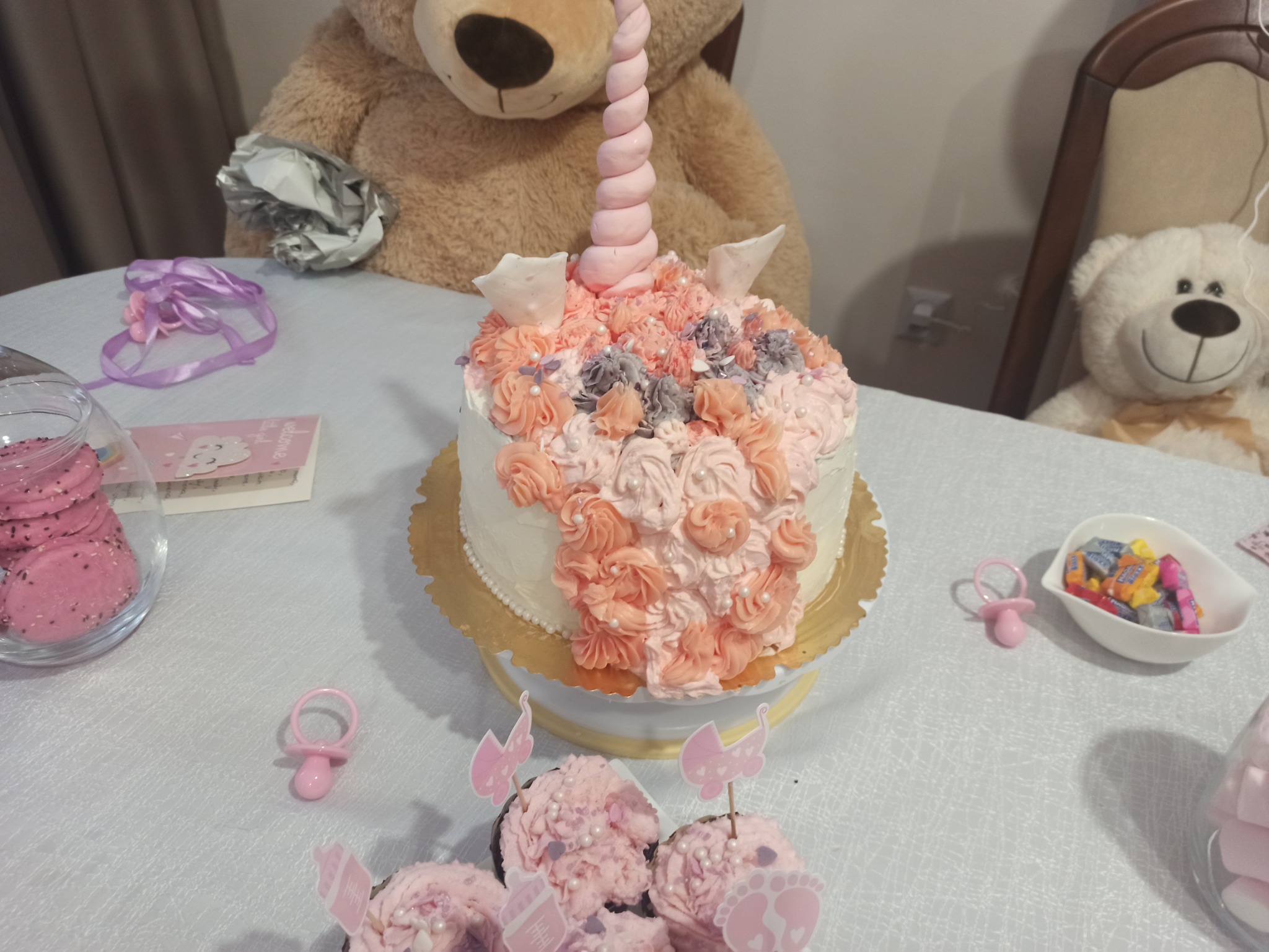Tort jednorożec z różowymi i szarymi kremowymi rozetami, otoczony rekwizytami dziecięcymi na stole, w tle pluszowe misie.