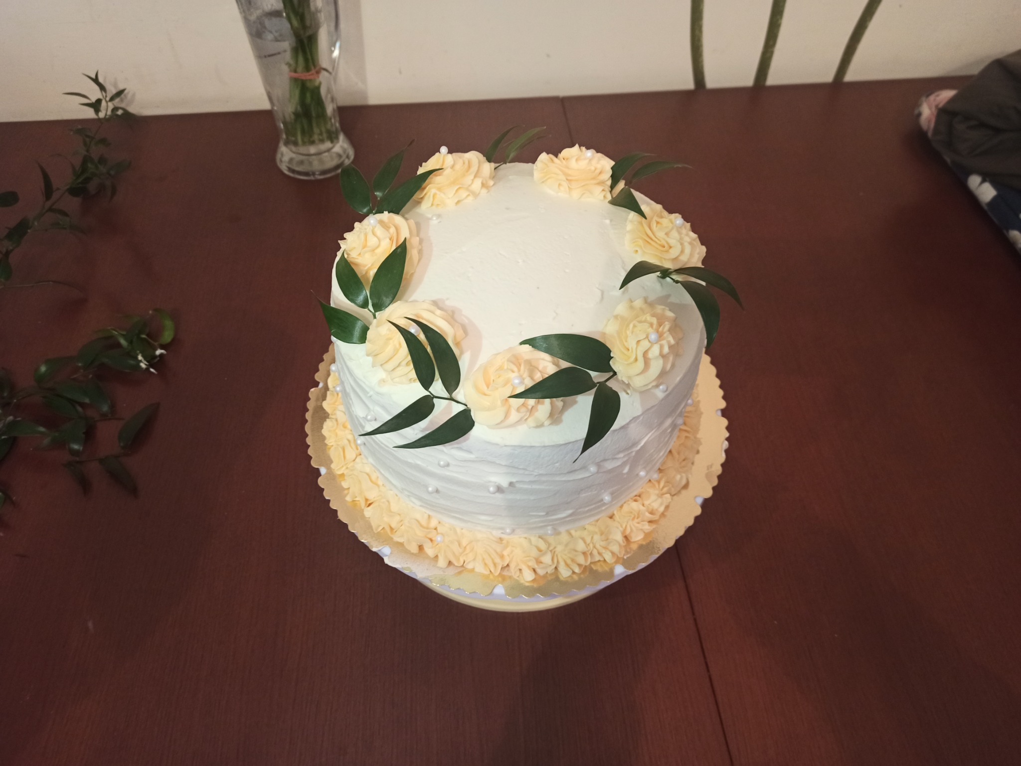 Dwupoziomowy tort z białym kremem, dekorowany kremowymi rozetkami, zielonymi listkami i perłowymi koralikami, umieszczony na złotej podkładce.