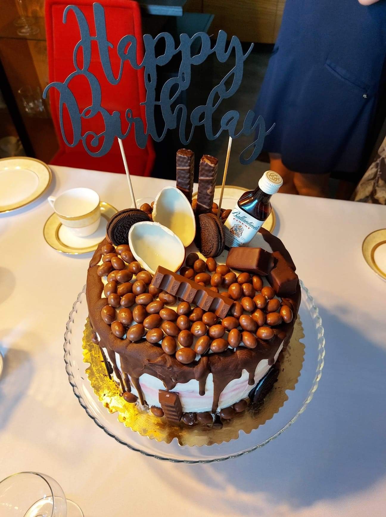 Udekorowany tort urodzinowy na szklanej paterze, z polewą czekoladową, drażami, batonikami, połówkami jajek z białej czekolady, ciasteczkami Oreo i miniaturową butelką whisky Ballantine's...