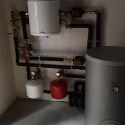 Bojlery, pompy obiegowe i rury tworzące system grzewczy w kotłowni, widoczne manometry i zawory.