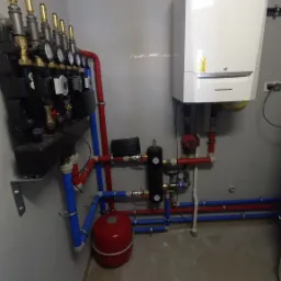 Nowoczesny, biały piec gazowy z podłączeniami hydraulicznymi w kolorach czerwonym i niebieskim, zamontowany na szarej ścianie w kotłowni. Widoczny rozdzielacz z manometrami, czerwony zbiornik...