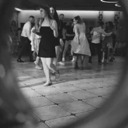 Czarno-białe zdjęcie z wesela: Młoda kobieta w czarnej sukience i białych tenisówkach tańczy na parkiecie w otoczeniu innych gości weselnych.
