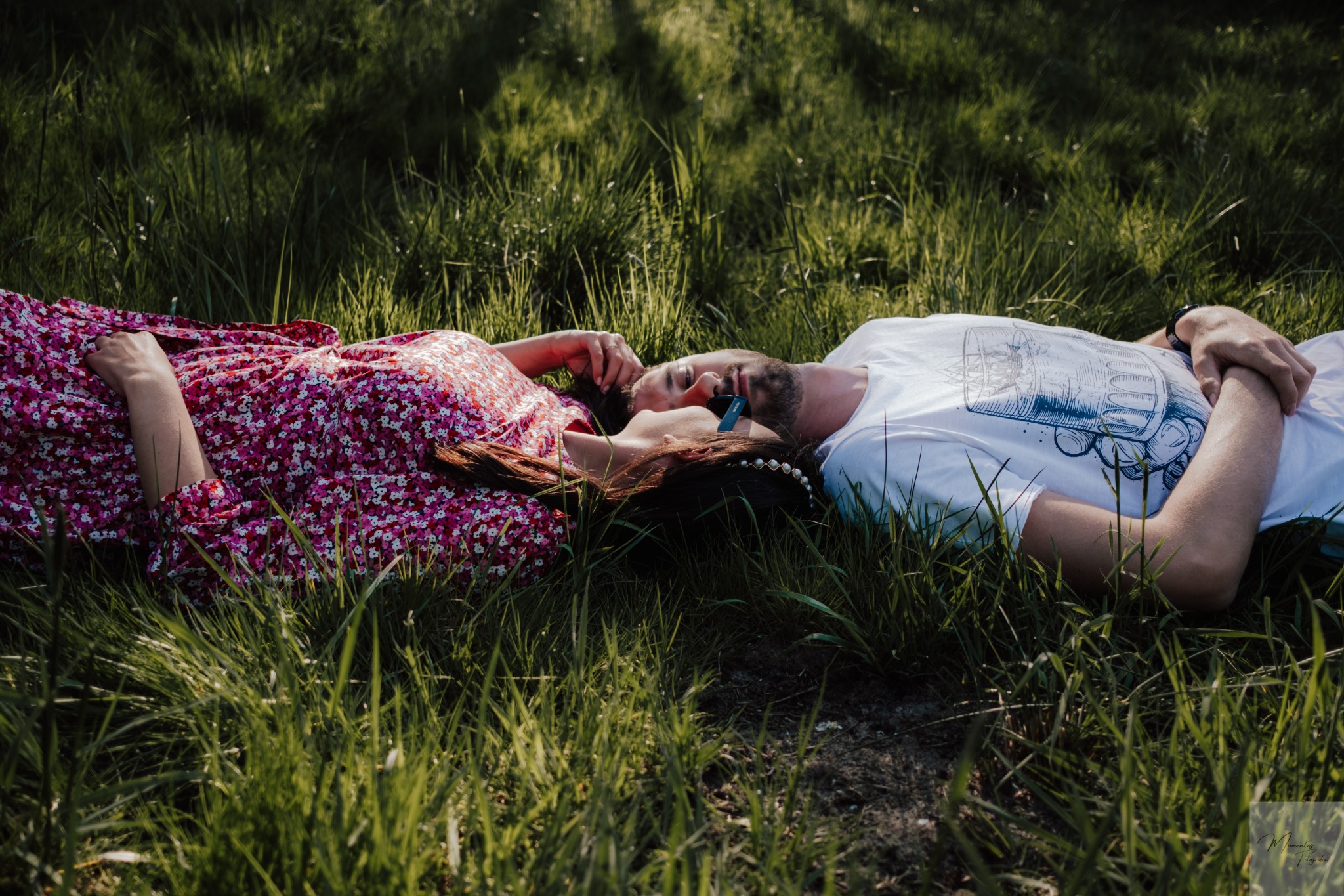 Para zakochanych leży na trawie, kobieta w kwiecistej sukience, mężczyzna w białym t-shircie z nadrukiem, zbliżenie twarzy, słoneczny dzień.