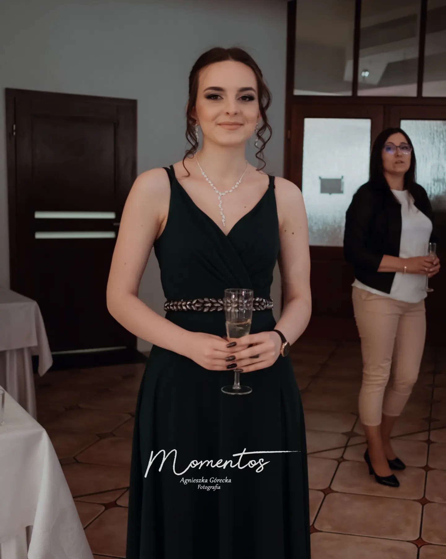 Elegancka kobieta w zielonej sukience trzyma kieliszek szampana, w tle druga kobieta i stół z białym obrusem. Ujęcie z uroczystości.