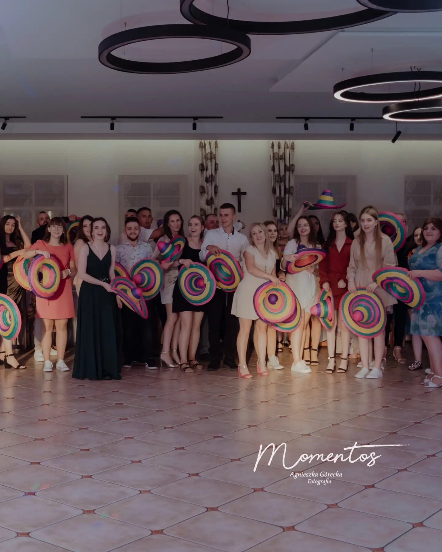 Grupa osób pozuje z kolorowymi sombrerami na tle sali z parkietem i nowoczesnymi lampami sufitowymi. W tle widoczne okna z zasłonami i krzyż na ścianie. Na pierwszym planie logo fotografa.