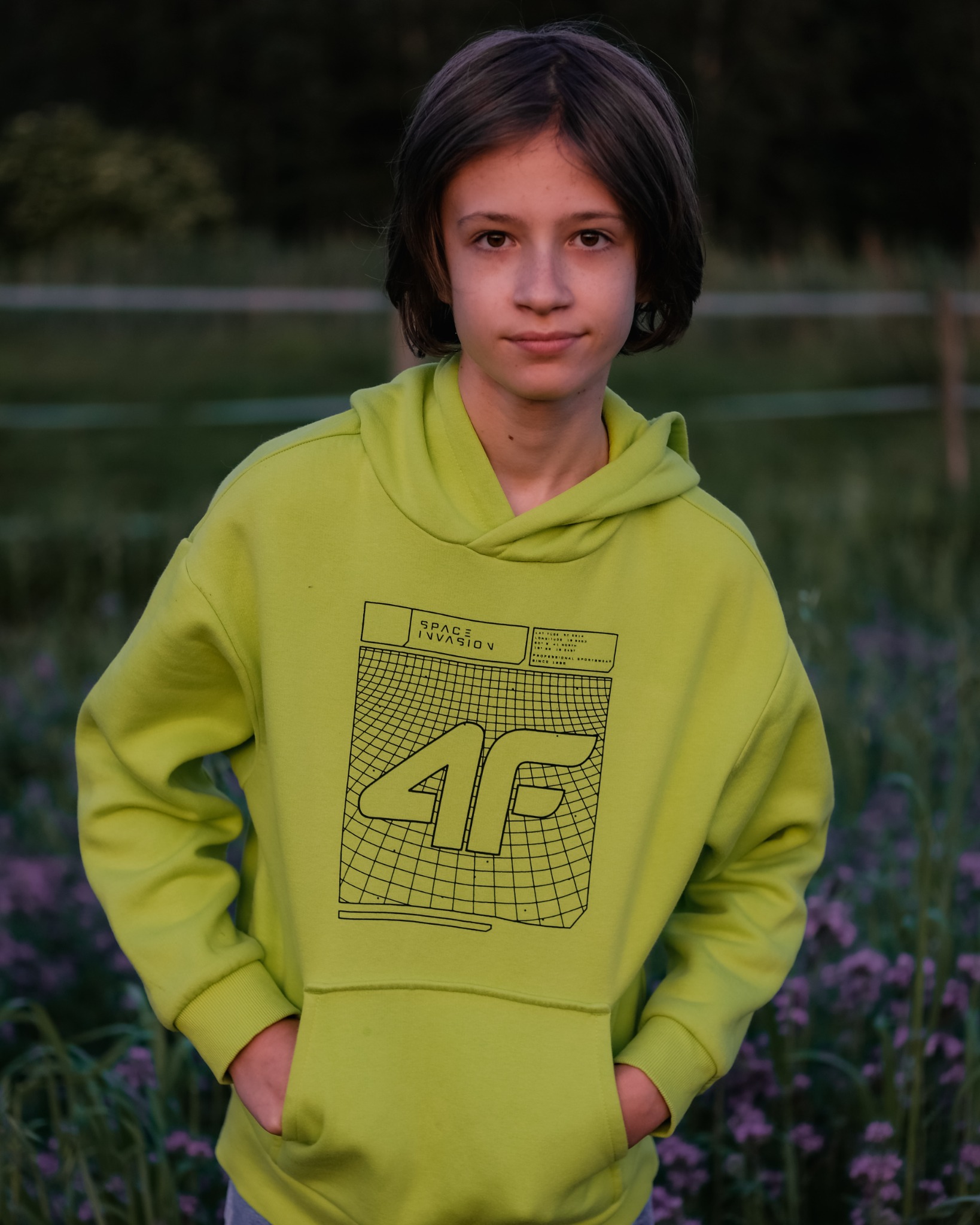 Portret nastolatka w limonkowym hoodie z nadrukiem 4F Space Invasion na tle zieleni i fioletowych kwiatów.