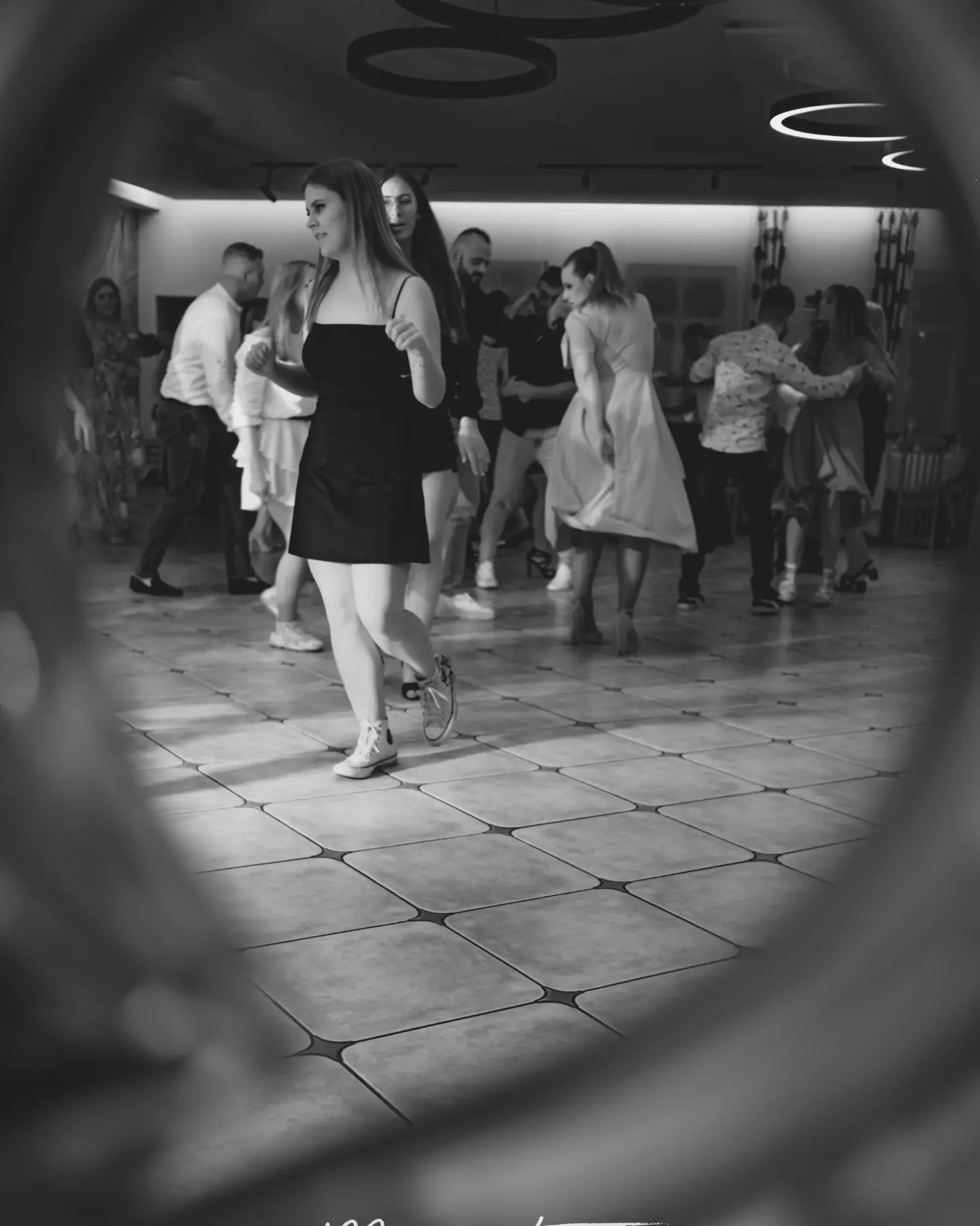 Czarno-białe zdjęcie z wesela: Młoda kobieta w czarnej sukience i białych tenisówkach tańczy na parkiecie w otoczeniu innych gości weselnych.