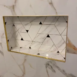 Wnęka w ścianie wyłożona białymi płytkami imitującymi marmur z geometrycznym wzorem w czarne trójkąty i złote linie, wykończona złotą listwą, zrealizowana w Żyrardowie.