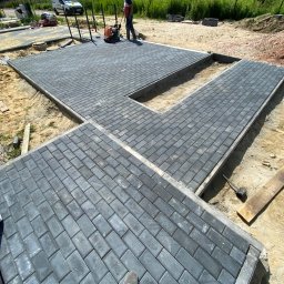 Pimpek - Układanie szarej kostki brukowej typu Holland na posesji, widoczna ubijarka wibracyjna w trakcie pracy, obrzeża betonowe, piasek i ziemia wokół, częściowo zakończony fragment nawierzchni.