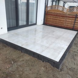 Pimpek - Jasne płyty ceramiczne 60x60 na tarasie, obrzeża z ciemnej kostki, widok na drewniany płot i okno tarasowe.
