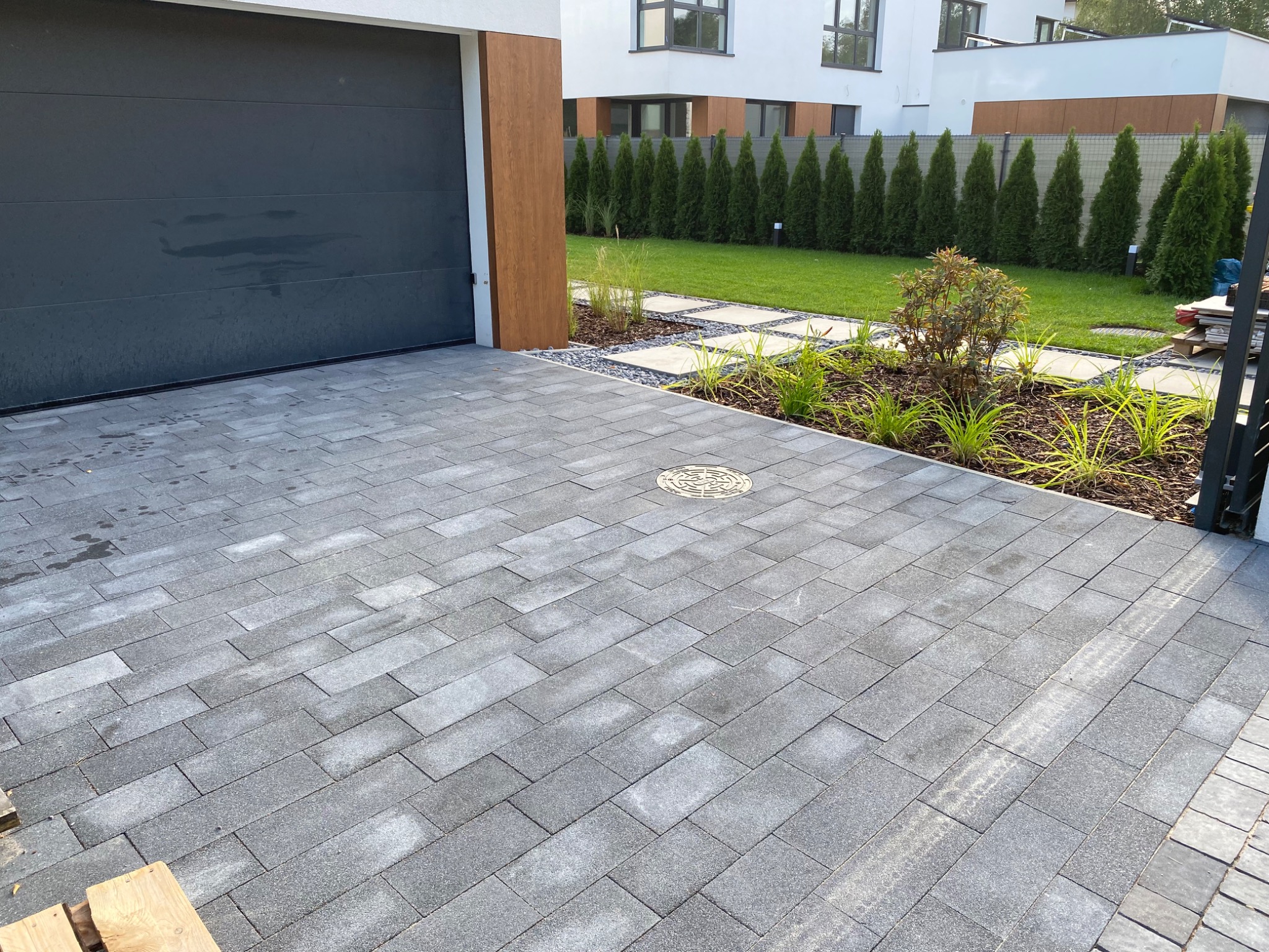 Ułożona szara kostka brukowa przed nowoczesnym domem z garażem, widoczna studzienka kanalizacyjna, trawnik z tujami w tle.