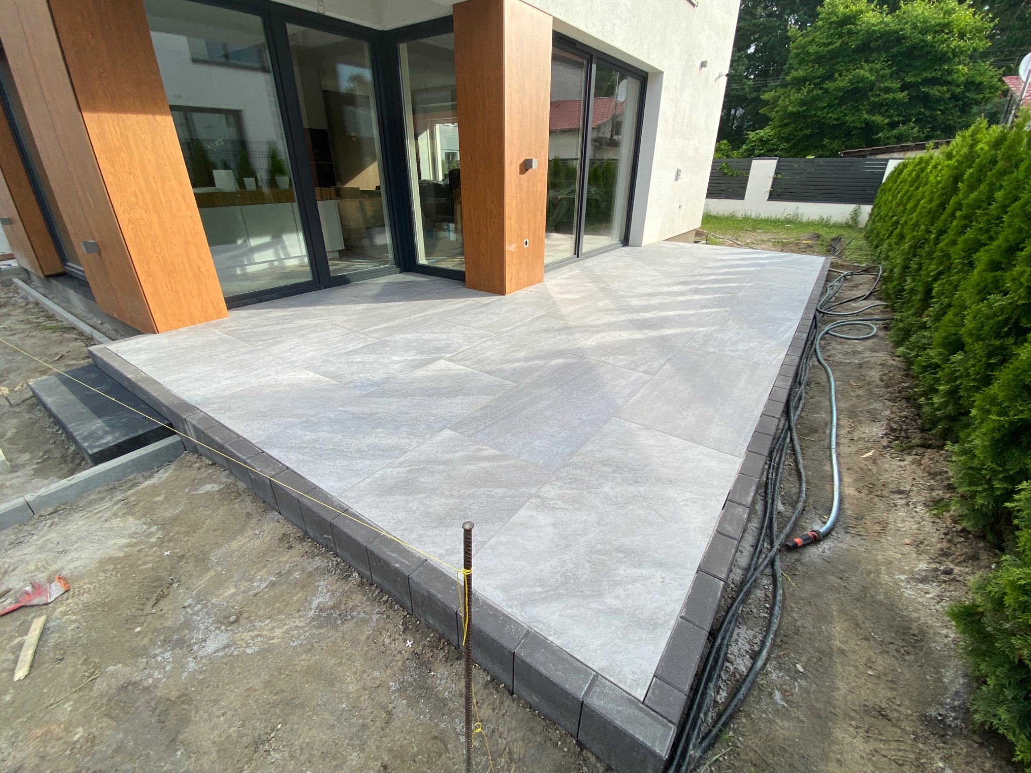 Nowoczesny taras z płyt ceramicznych Libet 60x120, z ciemnoszarą obwódką, w trakcie prac wykończeniowych, widoczne sznurki wyznaczające poziom i krawędzie, węże ogrodowe leżące obok
