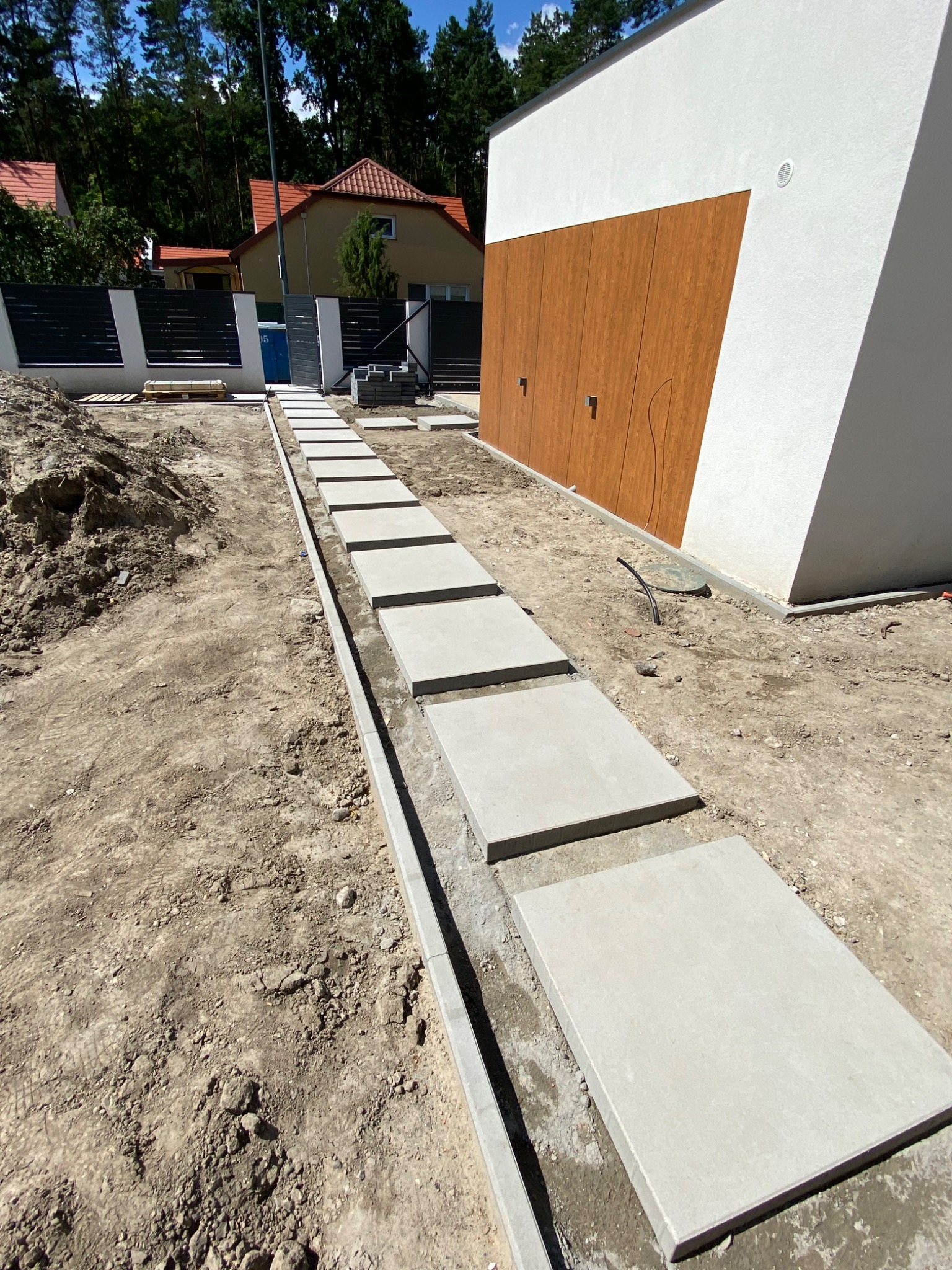 Ścieżka z płyt betonowych 80x80 cm, prawdopodobnie Libet Maxima, w trakcie układania, widoczna ziemia, obrzeża betonowe, nowoczesny dom z drewnianą elewacją w tle.