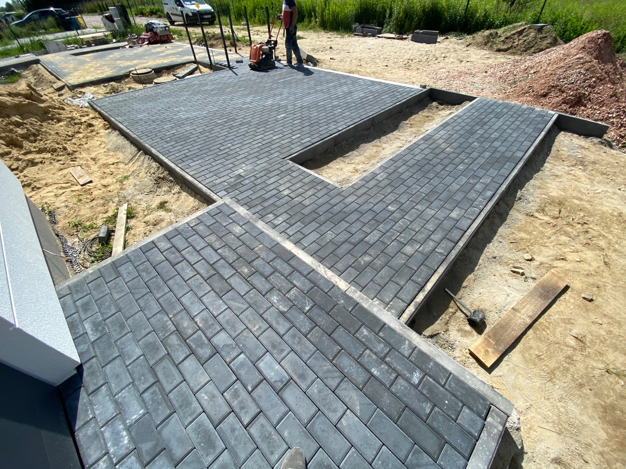 Układanie szarej kostki brukowej typu Holland na posesji, widoczna ubijarka wibracyjna w trakcie pracy, obrzeża betonowe, piasek i ziemia wokół, częściowo zakończony fragment nawierzchni.