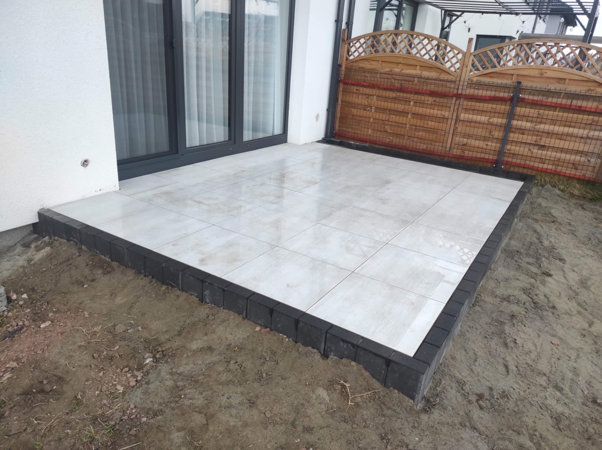 Jasne płyty ceramiczne 60x60 na tarasie, obrzeża z ciemnej kostki, widok na drewniany płot i okno tarasowe.