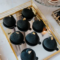 Czarne desery w kształcie serca z płatkami złota na lustrzanej tacy z ramą w kolorze złota, obok tiramisu w szklanych naczyniach.