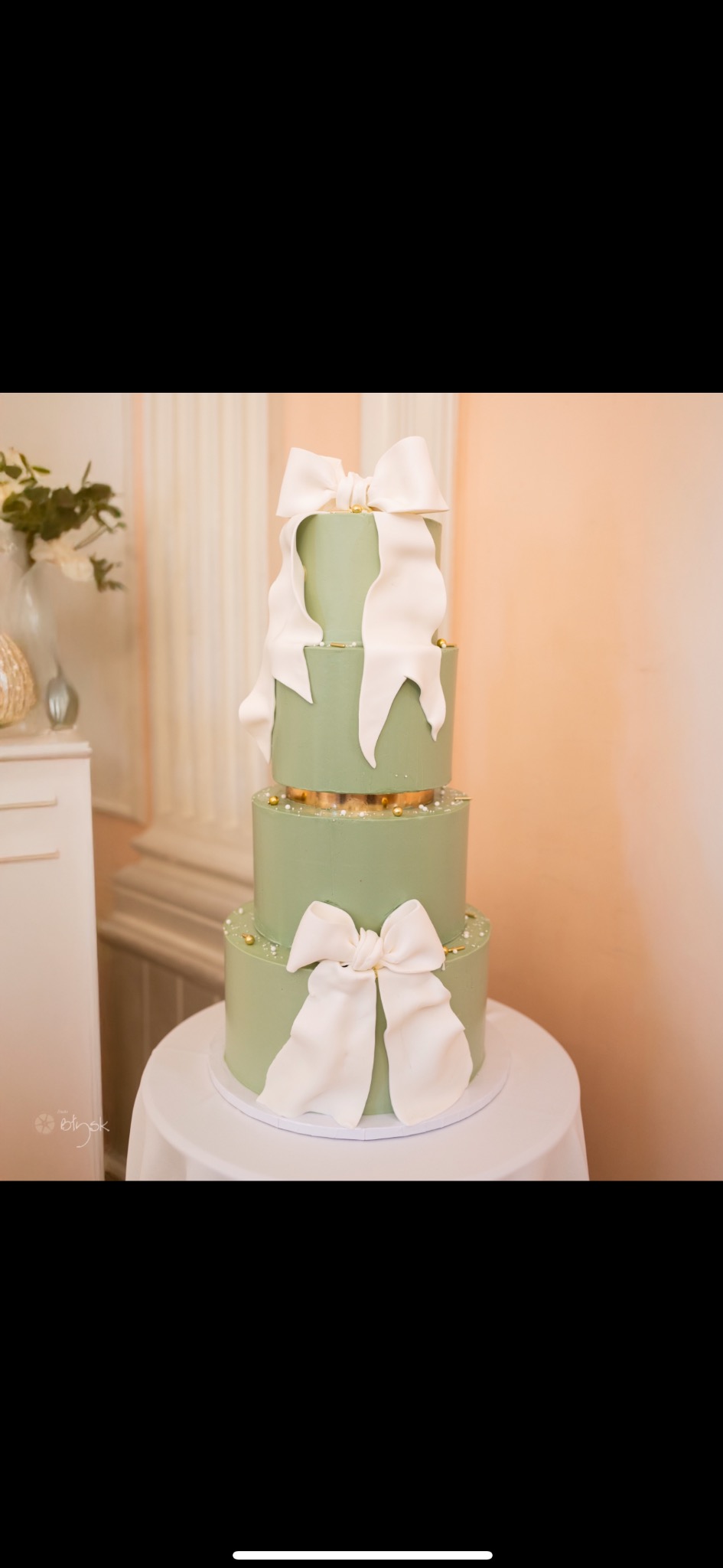 Trzypiętrowy tort w odcieniach zieleni, udekorowany dużymi, białymi kokardami i złotymi akcentami, na stole z białym obrusem, w tle kolumny i kwiaty.