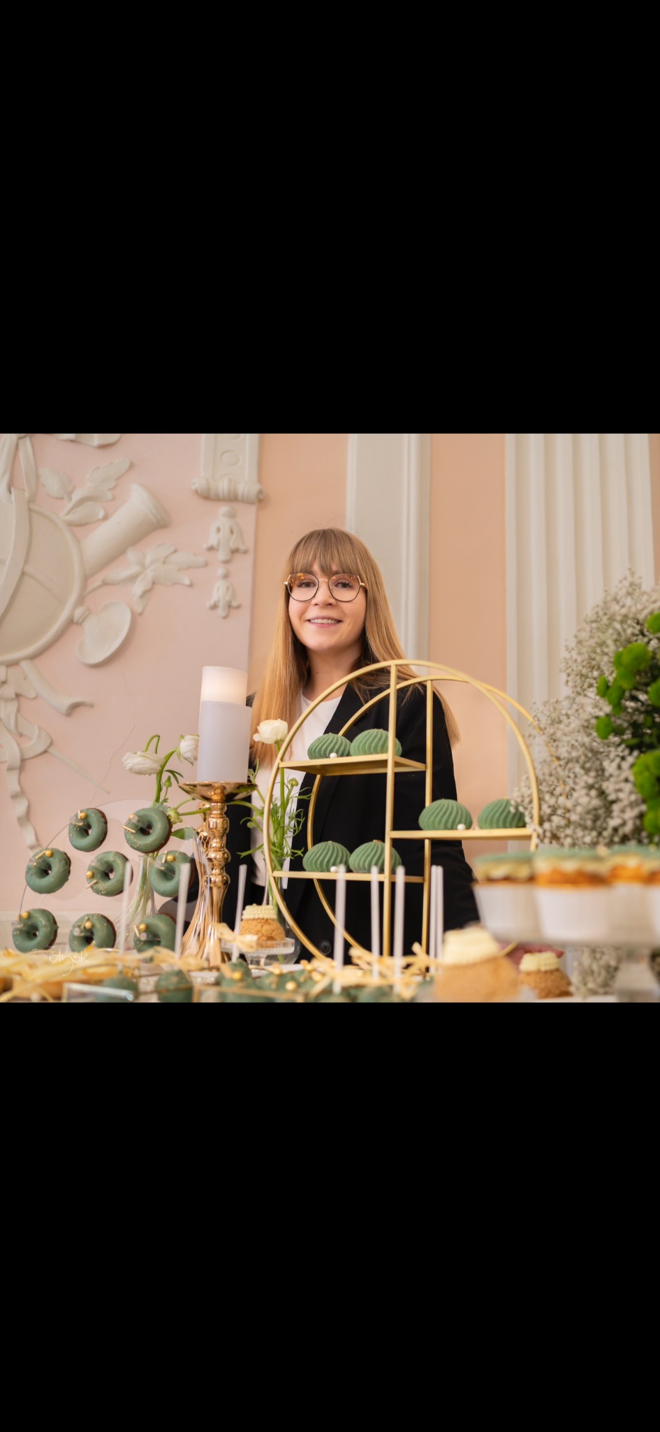 Uśmiechnięta blondynka za stołem z zielonymi donatami, ciastkami i świecami. Dekoracja na przyjęcie w pastelowych barwach z elementami złota.