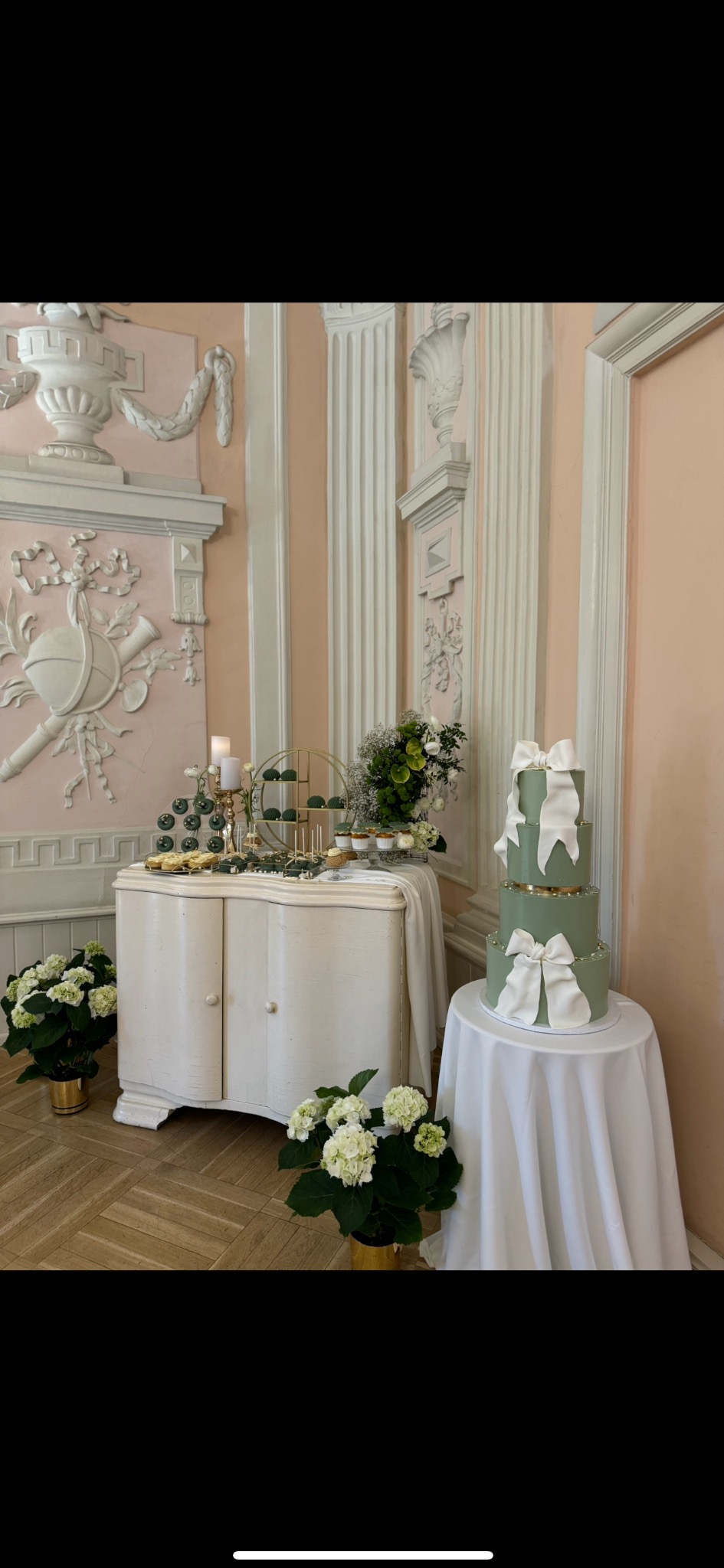 Elegancki, pistacjowy tort trzypiętrowy z kokardami, na stole z białym obrusem, w wystawnym wnętrzu. Obok bufet z deserami i kwiaty.