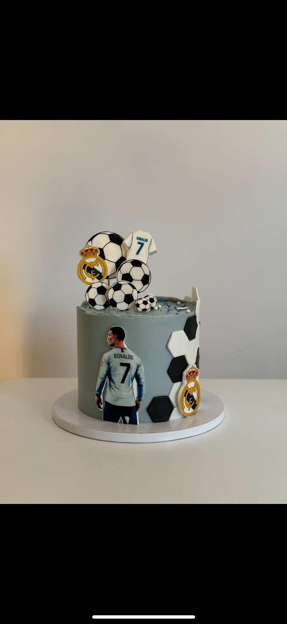 Szaro-biały tort z motywem piłki nożnej i Ronaldo, na białym tle. Dekoracje z piłek, koszulki i logo Realu Madryt. Widoczne heksagony na torcie.