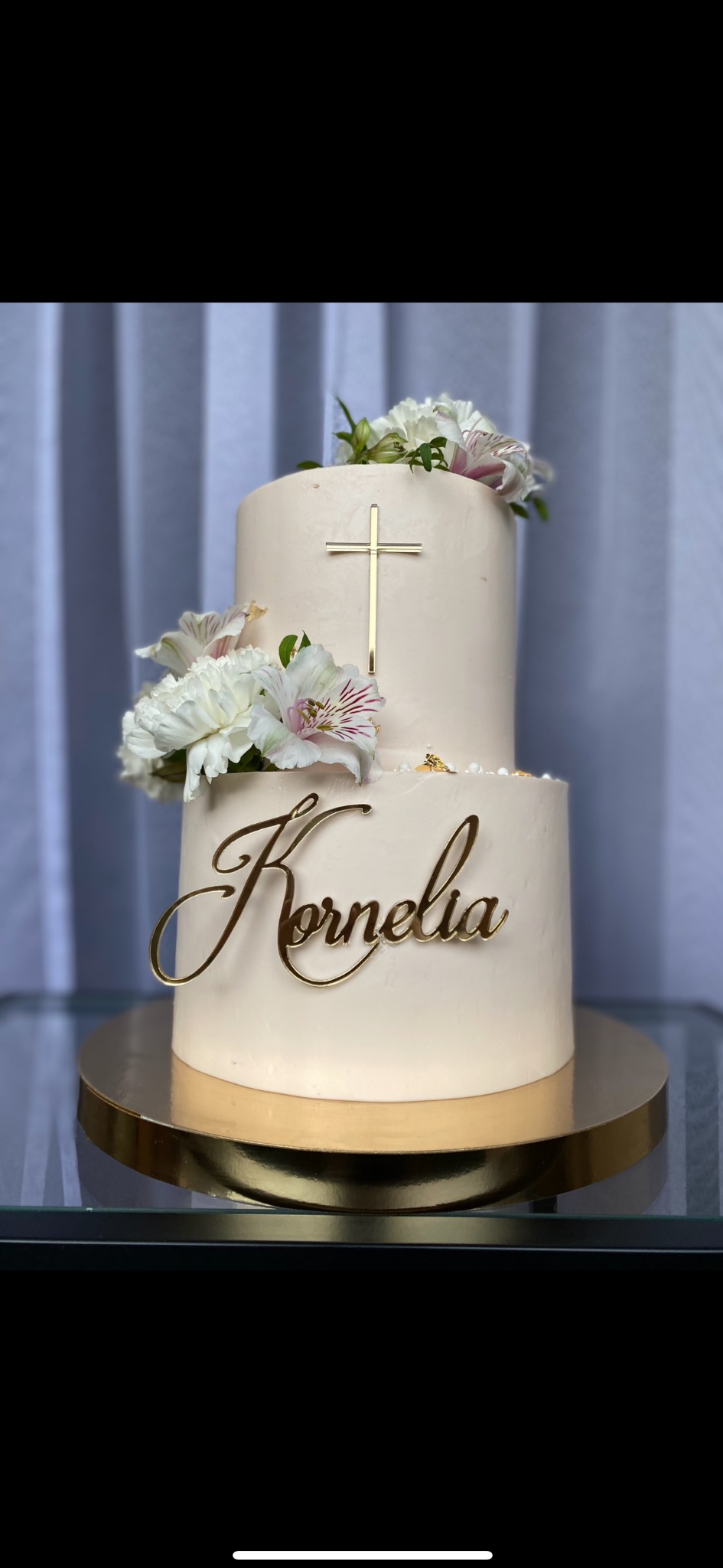 Dwupoziomowy tort w odcieniach kremu, zdobiony białymi kwiatami i złotym napisem 'Kornelia' oraz krzyżem, umieszczony na złotym podeście.